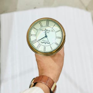 May include: A brass-coloured clock with a cream face and black Roman numerals. The clock face has the text "Royal Clocks Co. Est 1840 Scotland". The clock is held in a person's hand.