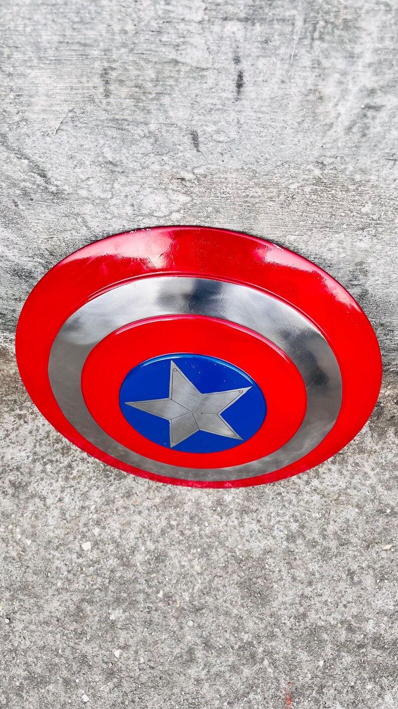 May include: A red, silver, and blue shield with a star emblem. The shield has a red outer ring, a silver middle ring, and a blue center with a silver star. The shield is a prop or collectible.
