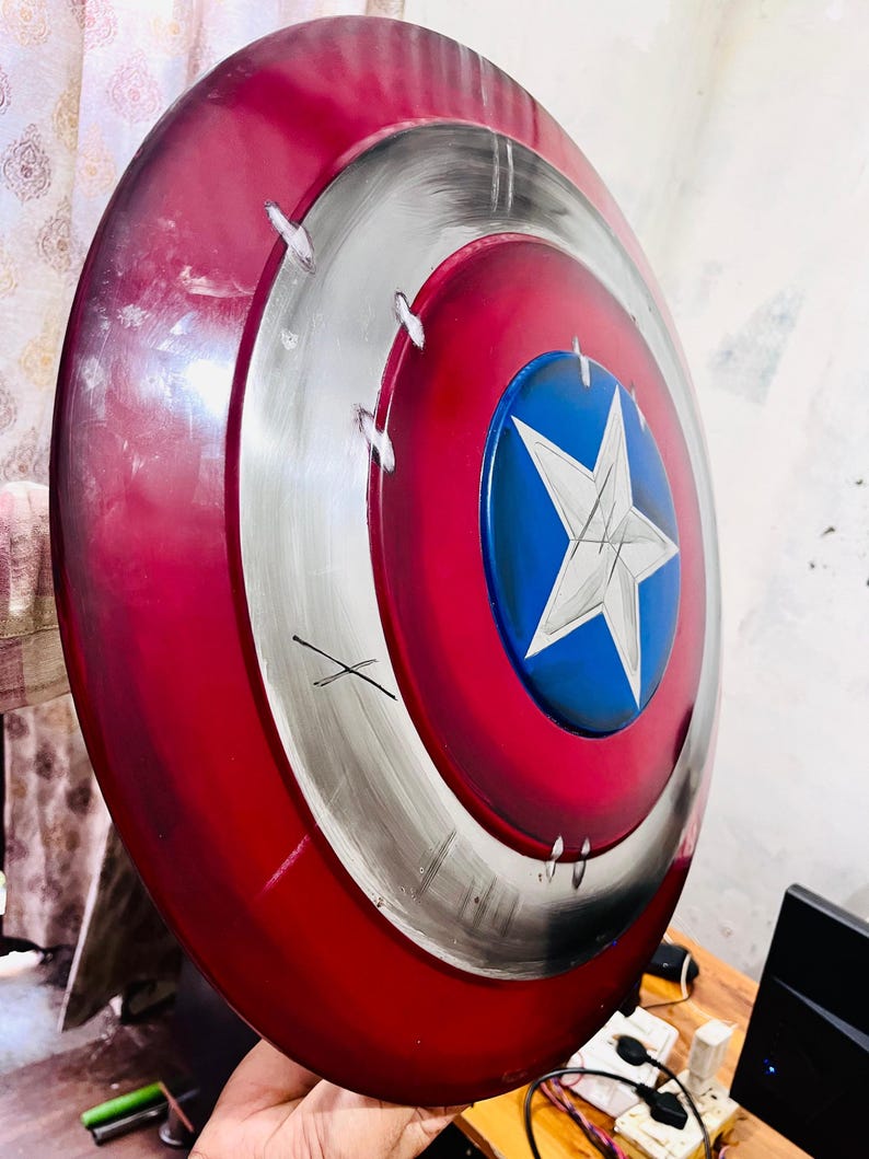 May include: A red, white, and blue shield with a star emblem. The shield has a circular design with a red outer ring, a silver middle ring, and a blue center with a white star. The shield has a distressed appearance.