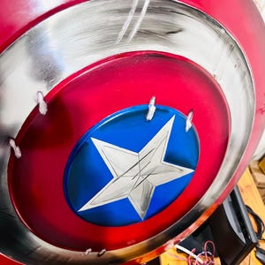 May include: A red, white, and blue shield with a star emblem. The shield has a circular design with a red outer ring, a silver middle ring, and a blue center with a white star. The shield has a distressed, battle-worn appearance.