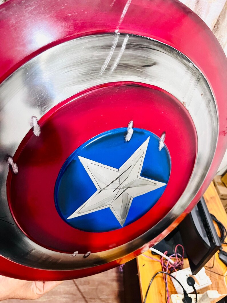 May include: A red, white, and blue shield with a star emblem. The shield has a circular design with a red outer ring, a silver middle ring, and a blue center with a white star. The shield has a distressed, battle-worn appearance.