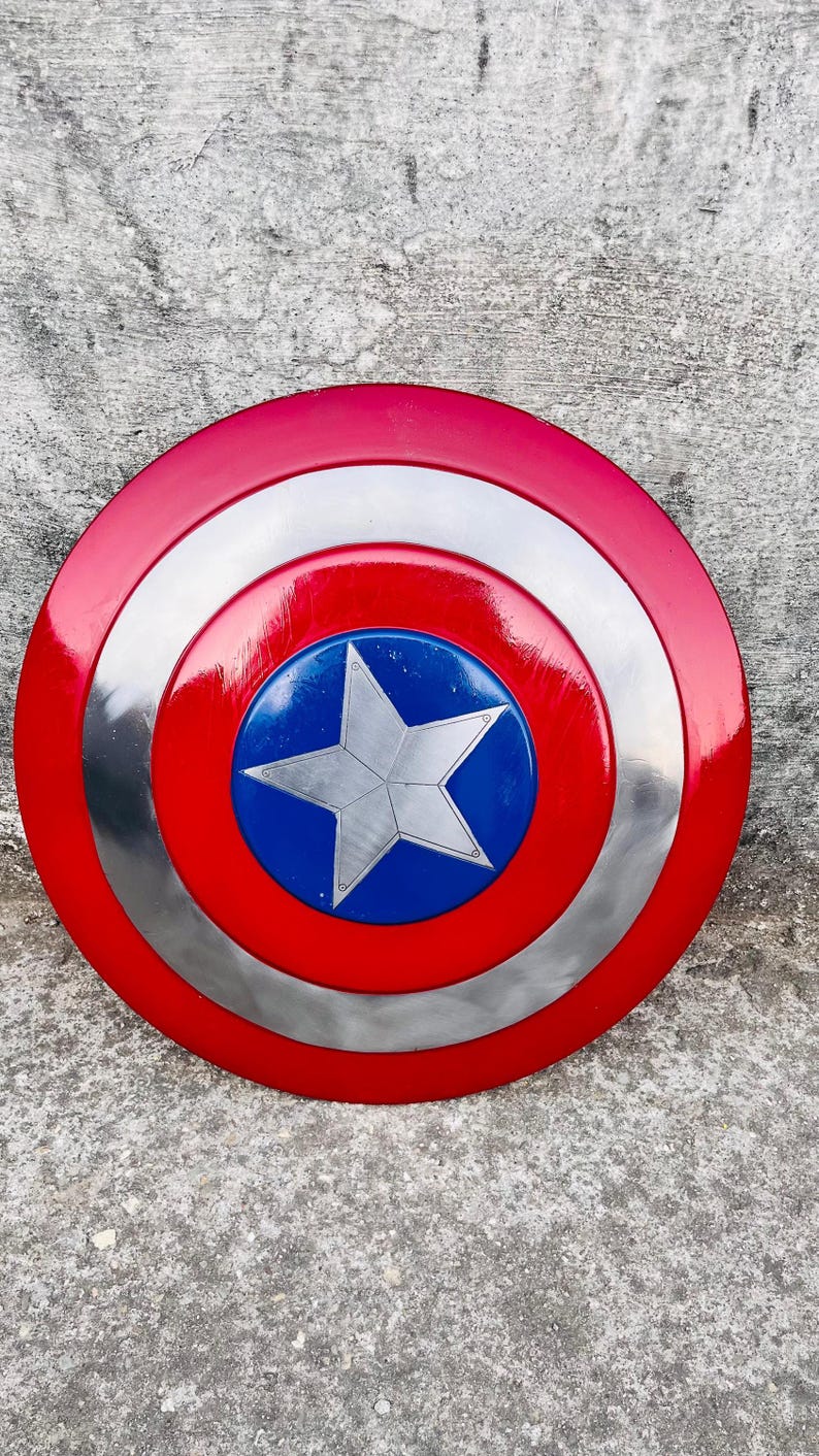 May include: A red, white, and blue shield with a silver star in the center. The shield has a circular design with concentric rings of red, silver, and blue. The shield is a prop or collectible.