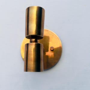 Op de afbeelding: Een messing wandlamp met een cilindrisch ontwerp. De lamp heeft een ronde achterplaat en twee cilindrische lichthouders. De afwerking is een warme, metallic goudkleur. De lamp is gemonteerd op een lichtblauwe muur.