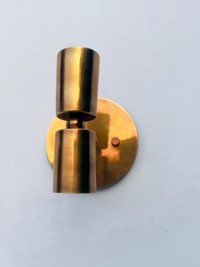 Op de afbeelding: Een messing wandlamp met een cilindrisch ontwerp. De lamp heeft een ronde achterplaat en twee cilindrische lichthouders. De afwerking is een warme, metallic goudkleur. De lamp is gemonteerd op een lichtblauwe muur.