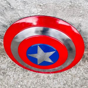 May include: A red, silver, and blue shield with a star emblem. The shield has a red outer ring, a silver middle ring, and a blue center with a silver star. The shield is a prop or collectible.