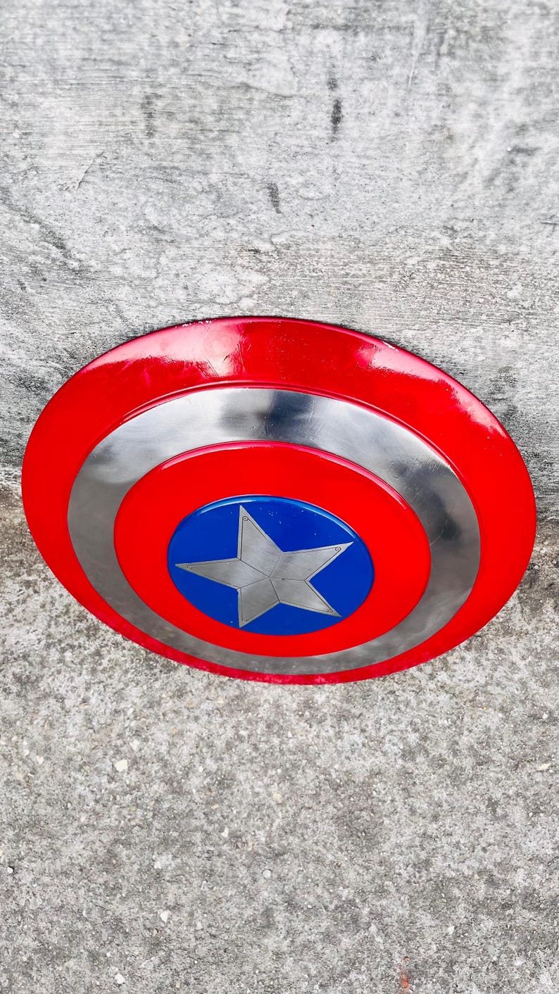 May include: A red, silver, and blue shield with a star emblem. The shield has a red outer ring, a silver middle ring, and a blue center with a silver star. The shield is a prop or collectible.