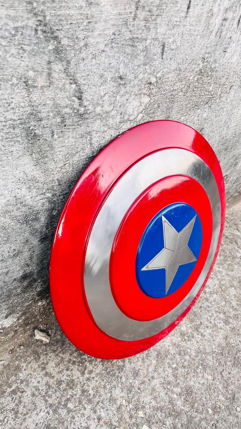 May include: A red, white, and blue Captain America shield. The shield has a red outer ring, a silver middle ring, and a blue center with a silver star. The shield is a circular shape.