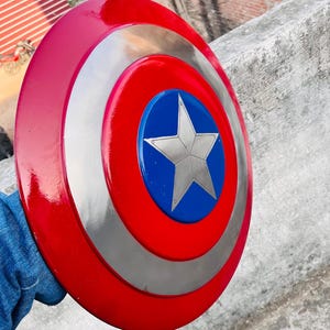 May include: A red, silver, and blue shield with a star in the center. The shield is round with a red outer ring, a silver middle ring, and a blue center with a silver star. A classic superhero accessory.