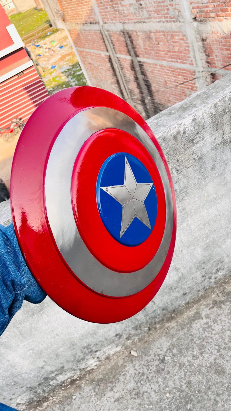 May include: A red, silver, and blue shield with a star in the center. The shield is round with a red outer ring, a silver middle ring, and a blue center with a silver star. A classic superhero accessory.