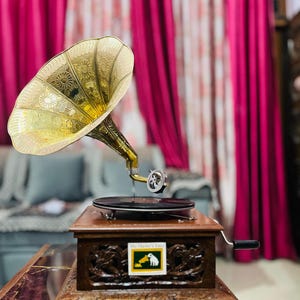 May include: A vintage gramophone with a gold-colored horn and a wooden base. The horn has intricate floral patterns. The base features carved details and a label that reads "His Master's Voice" with a dog and a gramophone. The gramophone is set on a marble-like surface.