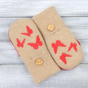 May include: A pair of beige mittens with red butterfly designs. The mittens have a red lining and are adorned with wooden buttons. The mittens are displayed on a light gray wooden surface.