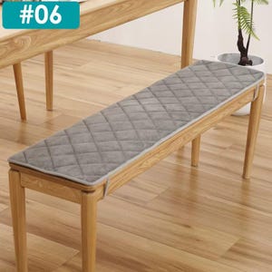 Puede incluir: Un cojín de banco acolchado gris con un patrón de diamantes sobre un banco de madera. El cojín tiene un borde gris claro. El banco es de madera clara. El texto "#06" está en un rectángulo turquesa en la esquina superior izquierda.