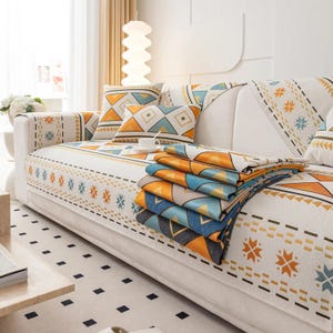 Könnte beinhalten: Ein weißes Sofa mit Kissen und einer Decke mit geometrischen Mustern in Orange, Blau und Gelb. Das Sofa steht in einem hellen Raum. Die Decke ist auf dem Sofa gefaltet.