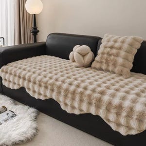 Puede incluir: Un sofá de cuero negro con una manta beige texturizada y cojines decorativos a juego. La manta y los cojines tienen un aspecto suave y afelpado. También hay un pequeño cojín decorativo redondo beige en el sofá.