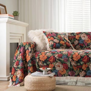 May include: A sofa adorned with a floral patterned cover and cushions. The cover displays a dark backdrop with vivid orange, red, and blue blooms. A white fringed trim and a fluffy white cushion provide texture. A small woven ottoman is positioned in front of the sofa.
