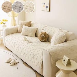 May include: A cream sofa with a textured, patterned cover and matching cushions. The sofa is accessorized with a small side table and a clock. The image also shows a rug and slippers. The sofa cover has a geometric design.