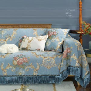 Puede incluir: Una toalla de sofá de chenilla azul claro con un estampado floral y flecos. La funda del sofá presenta un diseño de rosas y detalles dorados ornamentados. La imagen también muestra cojines decorativos y un gato blanco.