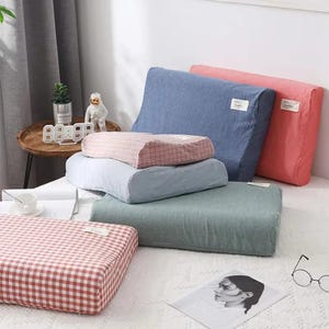 May include: A collection of pillows in various colors and patterns. The pillows include solid blue, pink, and green rectangular pillows, and a pink and white checkered pillow. Some pillows have small white tags.