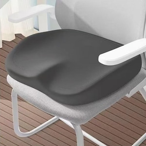 May include: A dark gray ergonomic seat cushion with a curved design, placed on a white and gray office chair. The cushion is designed to provide support and comfort. The chair has white armrests and legs.