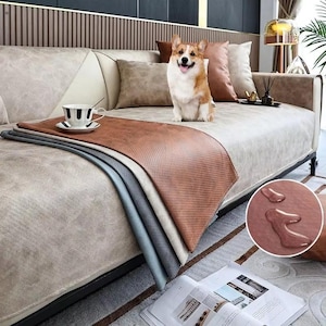 Könnte beinhalten: Ein beiges Sofa mit einer braunen und beigen Decke. Ein Corgi-Hund sitzt auf dem Sofa. Eine schwarz-weiße Tasse und Untertasse befinden sich auf dem Überwurf. Der Überwurf besteht aus wasserabweisendem Material.