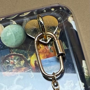 Pode incluir: Close-up de um mosquetão dourado com um pingente em forma de coração. O mosquetão está preso a uma capa transparente com um design floral. O mosquetão tem um portão com mola e uma pequena corrente.