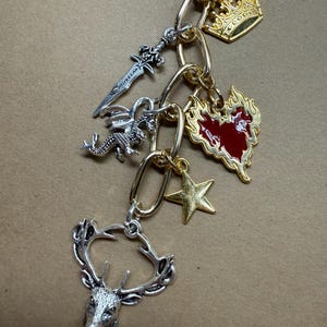 Puede incluir: Una pulsera de dijes dorada y plateada con varios colgantes. Los dijes incluyen una corona, una espada, un dragón, una estrella, un corazón y una cabeza de ciervo. La pulsera tiene una cadena dorada y detalles plateados.