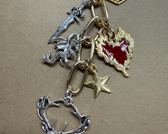 Könnte beinhalten: Ein gold- und silberfarbenes Charm-Armband mit verschiedenen Anhängern. Zu den Charms gehören eine Krone, ein Schwert, ein Drache, ein Stern, ein Herz und ein Hirschkopf. Das Armband hat eine goldene Kette und silberne Akzente.