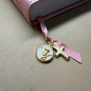 May include: A pink book with a matching ribbon bookmark. The bookmark features a gold cross charm and a round charm with a figure and star design. The book's cover is a light pink colour.