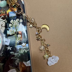 Puede incluir: Un dije de teléfono dorado con una cadena y dijes de temática celestial. Los dijes incluyen una luna creciente, estrellas, una nube y pequeñas cuentas. El dije está sujeto a una funda transparente para teléfono con un diseño de collage.