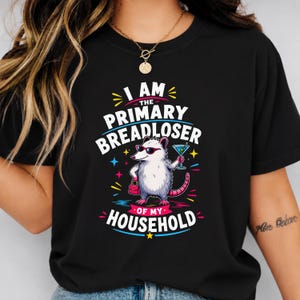 May include: A black t-shirt featuring a graphic of an opossum wearing sunglasses, holding a martini glass and a pink handbag. The text reads I AM THE PRIMARY BREADLOSER OF MY HOUSEHOLD in white, yellow, and pink lettering with colourful star accents.