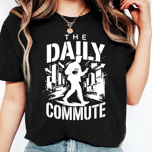 May include: Black t-shirt with a white graphic that reads "THE DAILY COMMUTE." The graphic depicts a person walking with a coffee cup and a phone, set against a city backdrop. The shirt is a casual wear item.