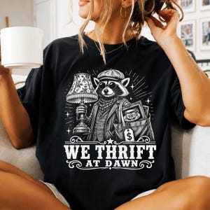 May include: A black t-shirt featuring a white graphic of a raccoon wearing a flat cap, sunglasses, and a scarf. The raccoon holds a vintage lamp and a tote bag containing a teacup. The text WE THRIFT AT DAWN is printed below the illustration.