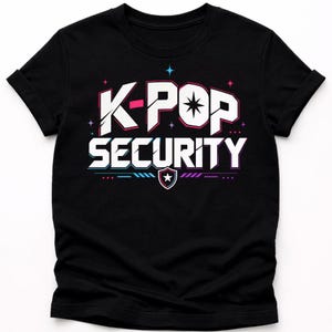以下が含まれることがあります： 白、ピンク、青で「K-POP SECURITY」の文字が入った黒いTシャツ。デザインには星と盾のエンブレムが含まれています。このシャツは半袖のクルーネックです。