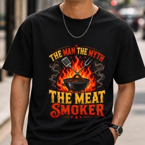 May include: Black t-shirt with a barbecue-themed graphic. The design features a grill, flames, and the text "THE MAN THE MYTH THE MEAT SMOKER" in a bold, vintage style. A spatula and fork are crossed above the grill.