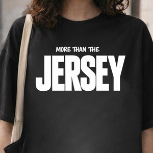 Puede incluir: Camiseta negra con el texto blanco "MORE THAN THE JERSEY". La camiseta es de cuello redondo. El texto está en negrita, con una fuente sans-serif.
