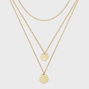 May include: A gold-colored layered necklace with three chains of varying lengths. The bottom chain features a textured, hammered-look pendant, while the middle chain has a smooth, circular pendant. The top chain is a simple, delicate chain.