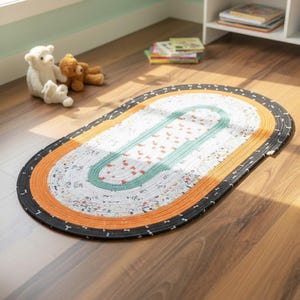Può includere: Tappeto ovale intrecciato con un design multicolore. Il tappeto presenta un centro bianco con piccoli motivi floreali, bordato da fasce turchesi, arancioni e nere. Il tappeto è posizionato su un pavimento in legno in una stanza con giocattoli.