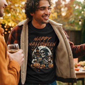 Op de afbeelding: Zwart T-shirt met een Halloween-afbeelding. Het ontwerp toont een schedel in een capuchon, twee heksen, pompoenen en een kasteel, met de woorden "Happy Halloween" in oranje.
