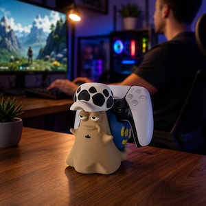 Op de afbeelding: Een beige slakvormige controllerstandaard met een witte hoed met zwarte stippen, een blauwe schelp met gele accenten en een klein zwart kroonsymbool op de borst, ontworpen om een wit-zwarte videogamecontroller stevig op zijn plek te houden.