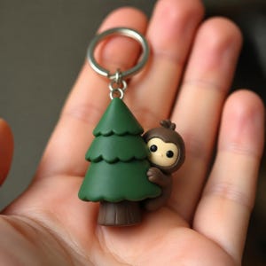 May include: A keychain featuring a small, brown, cartoon-style creature with large black eyes peeking from behind a dark green, tiered pine tree. The charm is attached to a circular silver metal split ring, creating a compact accessory.