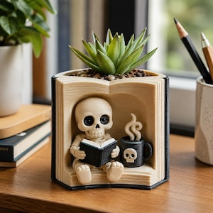May include: A beige planter shaped like an open book with a green succulent on top. Inside the book cavity, a small beige skeleton figure sits reading a black book next to a black mug with a white skull icon and white steam rising from the top.