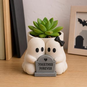 Peut inclure: Un pot de fleurs cylindrique blanc cassé avec deux figurines de fantômes blancs tenant une pierre tombale grise portant l'inscription TOGETHER FOREVER et un petit cœur. Une plante succulente verte est plantée avec un nœud noir.
