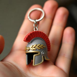 May include: A miniature grey Roman-style helmet keychain featuring a prominent red crest and a gold-colored laurel wreath detail across the front. The helmet is attached to a standard silver-toned metal split ring by a short chain link.