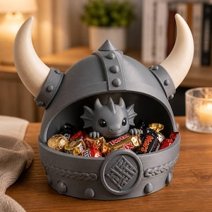 May include: A gray, bowl-shaped container designed like a Viking helmet with two large, curved, cream-colored horns. A small, gray dragon figure sits inside the bowl, which is filled with various individually wrapped chocolate candies.