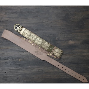 May include: A light brown leather belt with a decorative metal buckle and square metal plates. The metal plates have embossed designs. The belt is laid out on a dark wooden surface. The belt has a series of holes for adjustment.