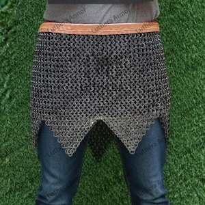 May include: A chainmail skirt with a brown leather belt. The skirt is made of small, interlocking metal rings. The bottom edge has a decorative, pointed design. The person is wearing blue jeans.