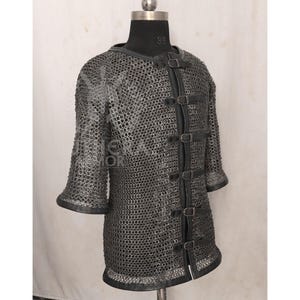 May include: A silver chainmail shirt with short sleeves and a leather trim. The shirt features leather straps with buckles down the front. The garment is displayed on a black mannequin.