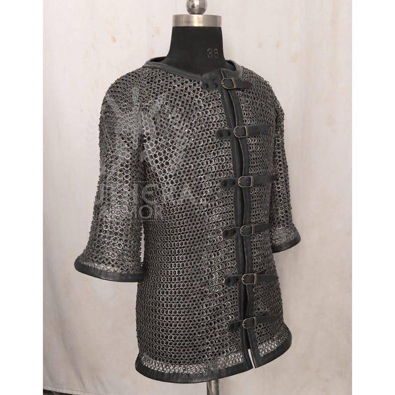 May include: A silver chainmail shirt with short sleeves and a leather trim. The shirt features leather straps with buckles down the front. The garment is displayed on a black mannequin.