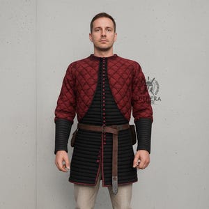 May include: A quilted burgundy and black medieval gambeson with a brown leather belt and a small pouch. The gambeson has long sleeves with black ribbed cuffs. The garment is buttoned up the front and has a red trim. The text "LUTHERA ARMOR" is visible.