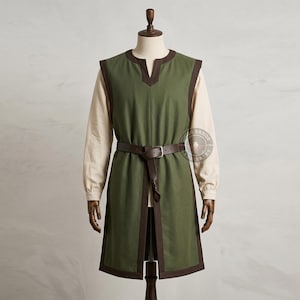 May include: A sleeveless olive green tunic with dark brown trim around the neckline, armholes, and hem, worn over a long-sleeved cream-colored shirt. A dark brown belt with a silver-toned buckle is cinched at the waist of the tunic.
